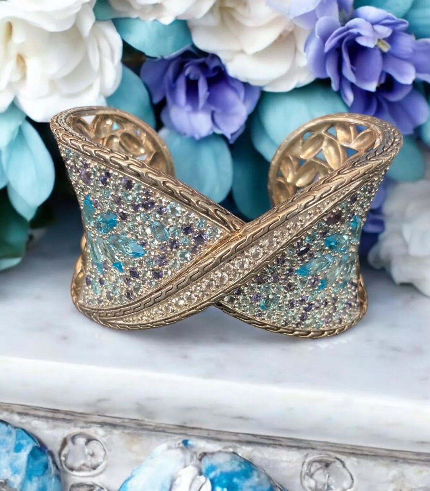 Gold cuff bracelet with blue and purple gemstone embellishments on marble surface with blue and white flowers