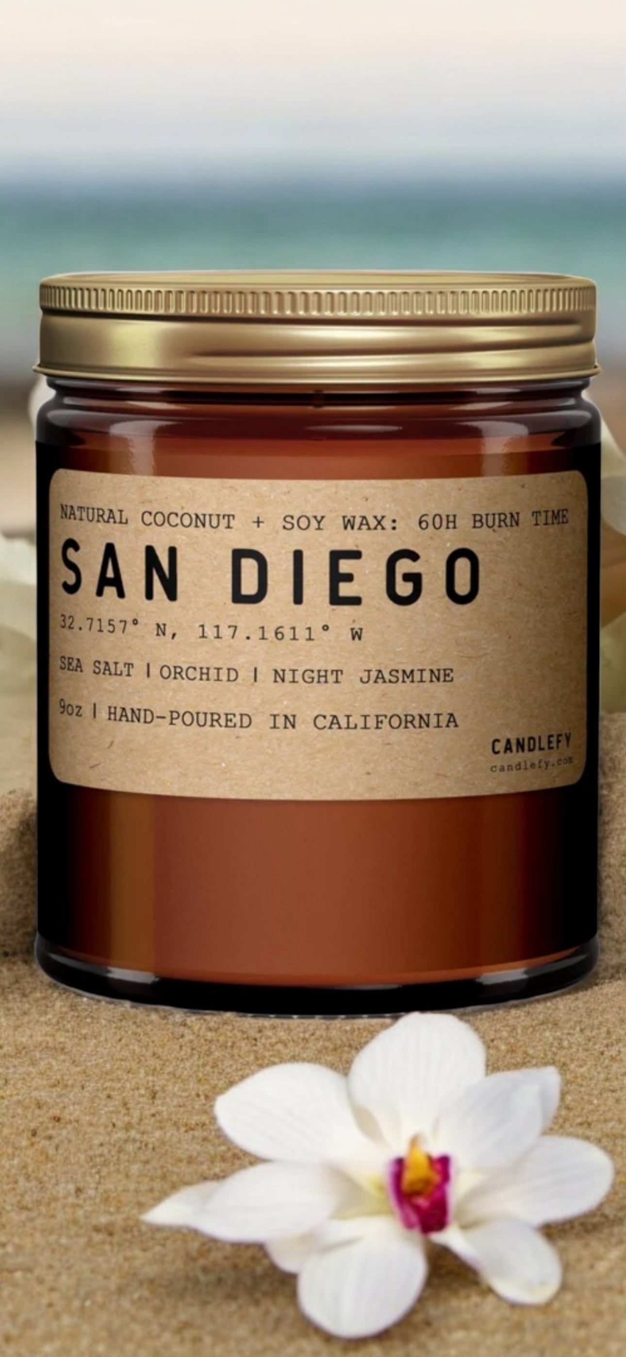 Amber glass candle jar labeled San Diego scent, natural coconut and soy wax, on sand with white orchid flower