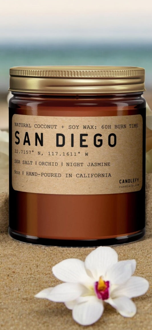 Amber glass candle jar labeled San Diego scent, natural coconut and soy wax, on sand with white orchid flower